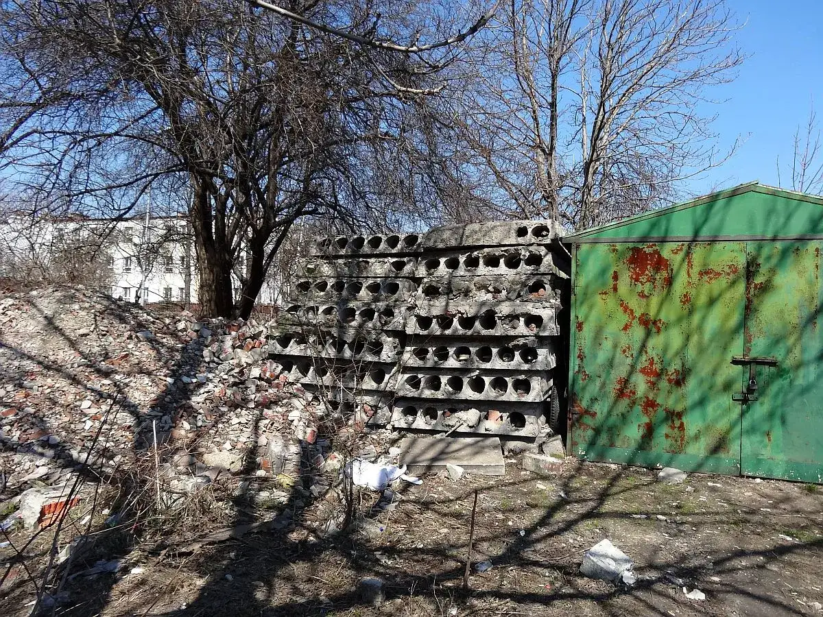 Stos płyt żerańskich, gruz i zielona, zardzewiała hala garażowa.