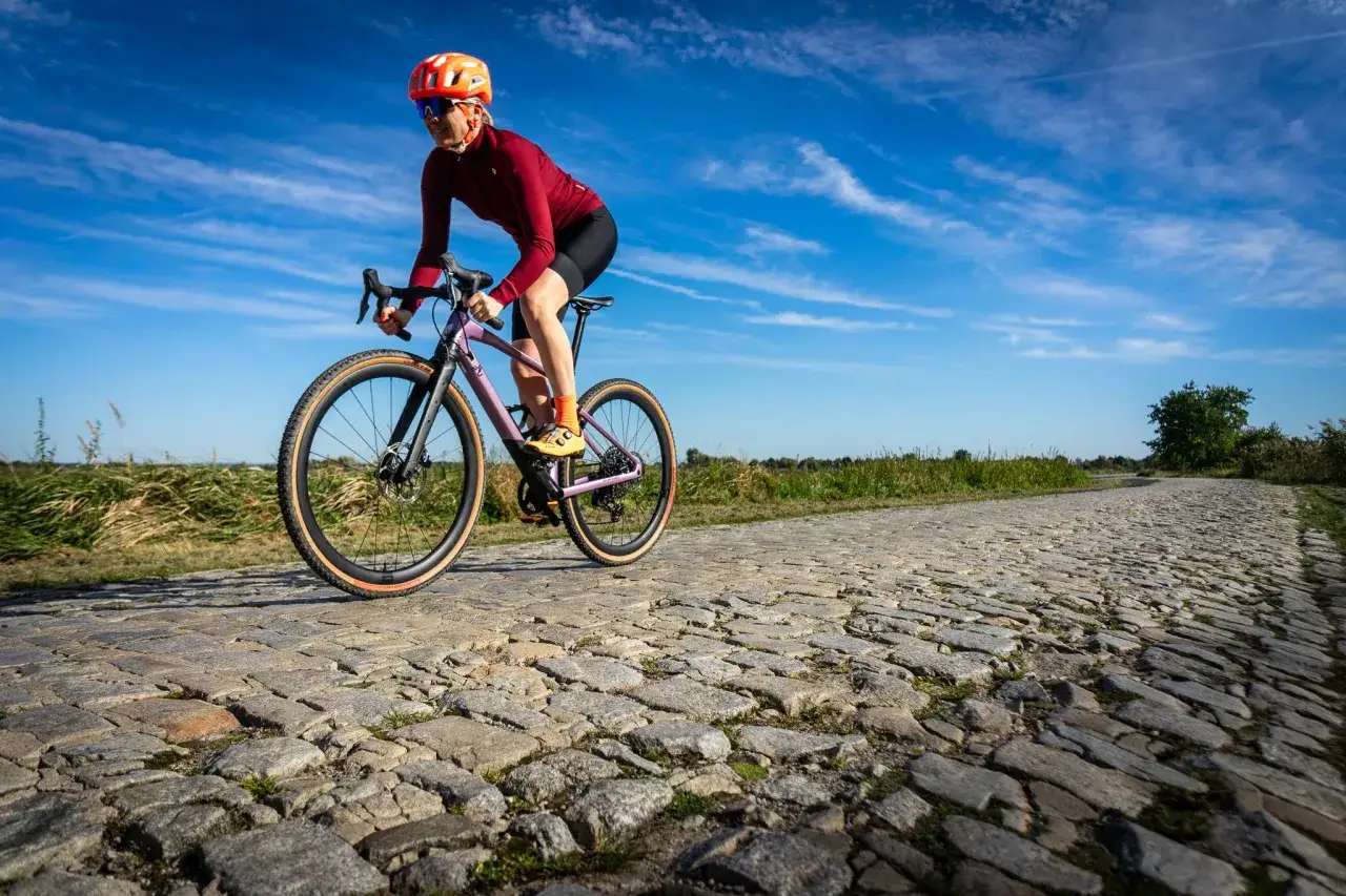 Kobieta w kasku i okularach jedzie na rowerze BMC URS LT po brukowanej drodze.