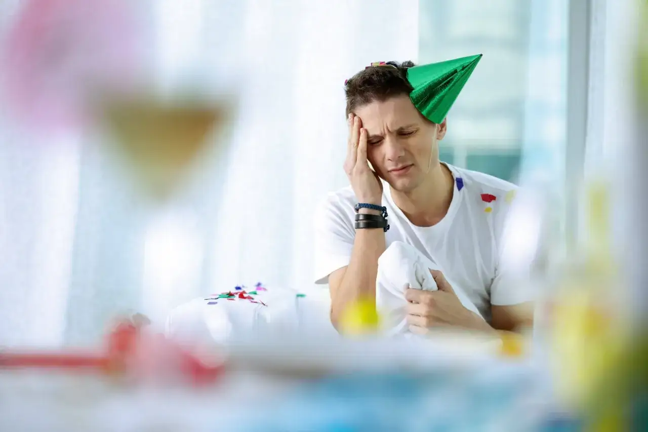 Mężczyzna z zielonym stożkiem na głowie, cierpiący na ból głowy po imprezie. Szuka sposobu, co na ból głowy na kacu.