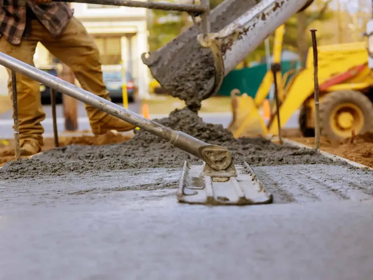 Pracownik wylewa beton, wyrównując go narzędziem. W tle koparka. Beton można lać do ok. 30°C.