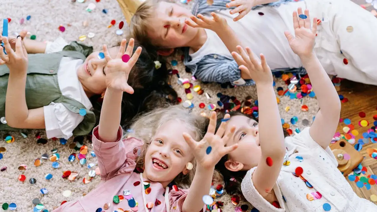 Dzieci leżą na dywanie wśród kolorowych konfetti, unosząc ręce w górę.