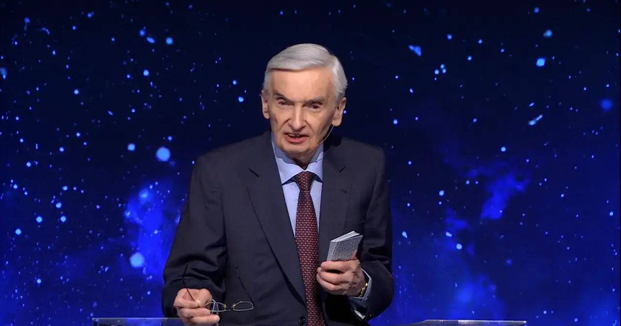 Stars twinkle behind a man in a suit, holding glasses and a small book. He looks like he's been on "Jeden z dziesięciu" for a long time.