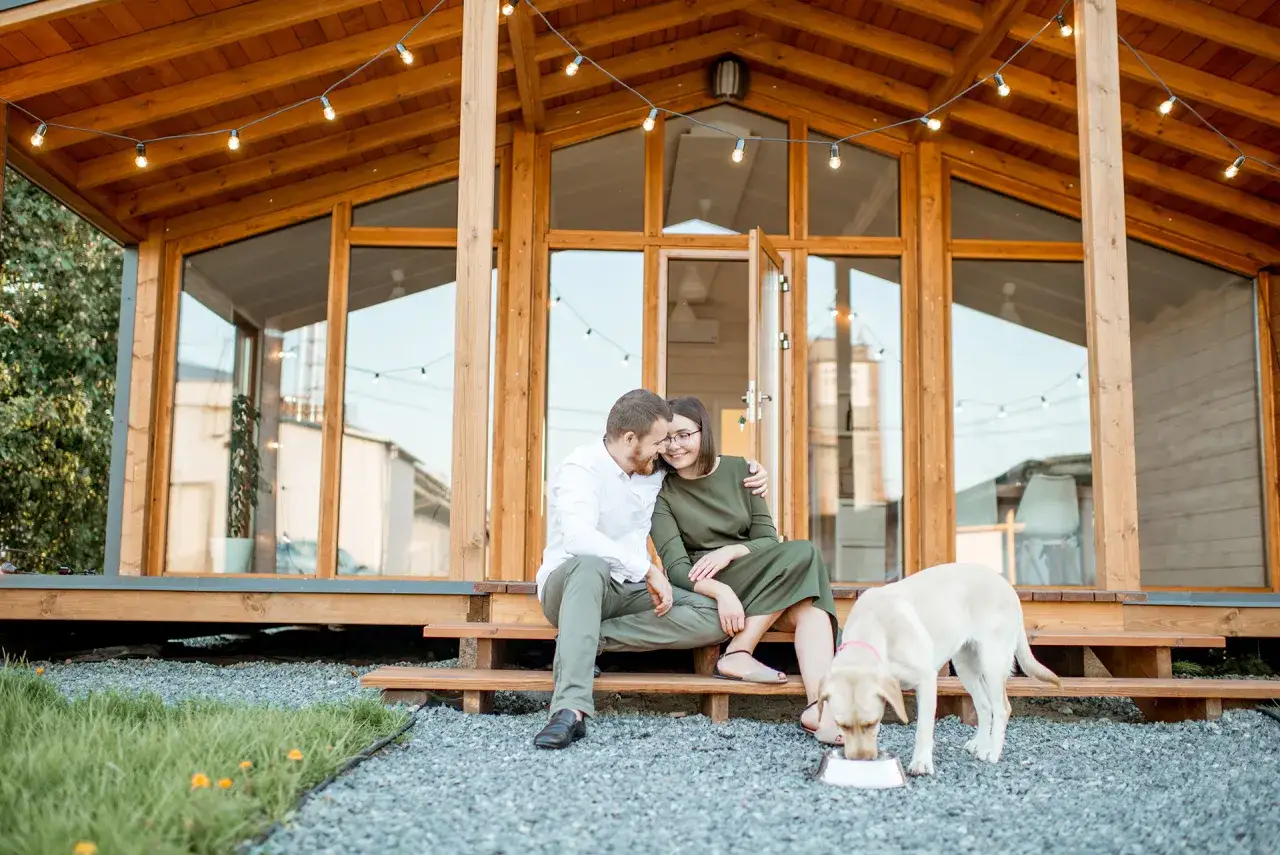 Para zakochanych siedzi na schodkach przed drewnianym domkiem letniskowym na zgłoszenie. Pies pije wodę z miski.