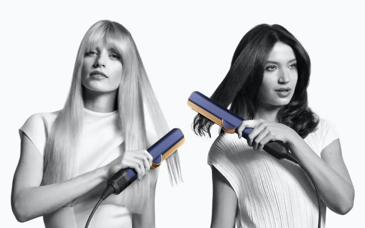 Dwie kobiety stylizujące włosy prostownicą Dyson Airstrait w kolorze granatowo-złotym.