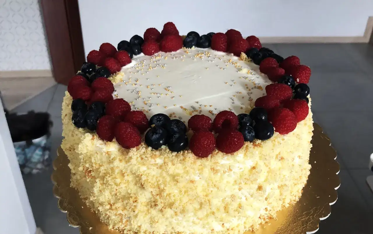 Pyszny tort śmietankowo-malinowy z mascarpone, ozdobiony świeżymi malinami i borówkami, posypany złotymi i srebrnymi kuleczkami.