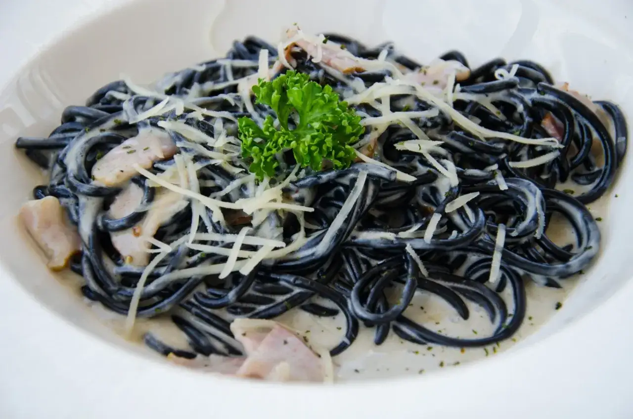 Czarny makaron spaghetti w kremowym sosie, posypany startym parmezanem i udekorowany gałązką natki pietruszki.