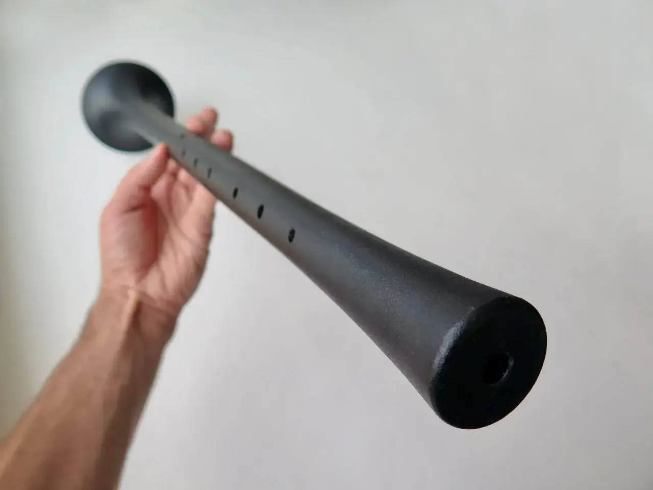 Dłoń trzyma czarną, podłużną piszczałkę podobną do oboju, z otworami na palce i rozszerzonym końcem.