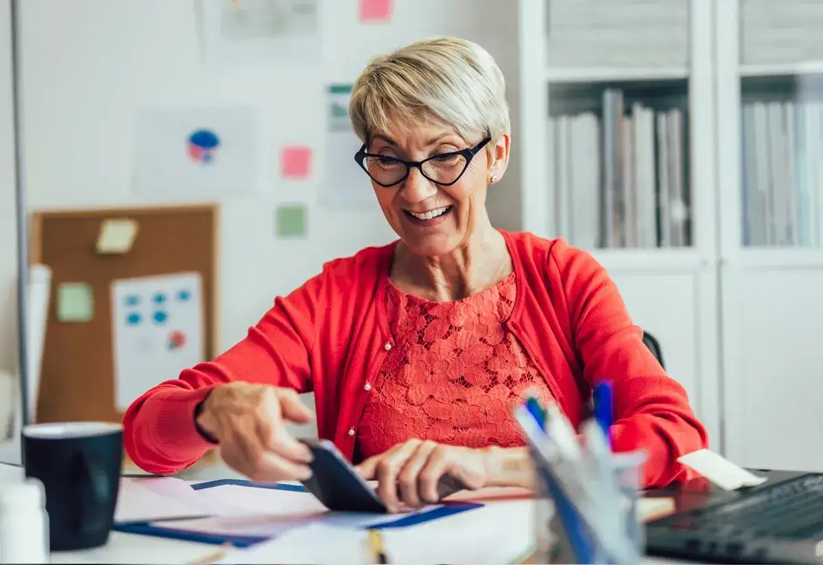 Uśmiechnięta starsza pani z okularami, pracująca przy biurku, sprawdza coś na telefonie. Zastanawia się, ile można dorobić na emeryturze.