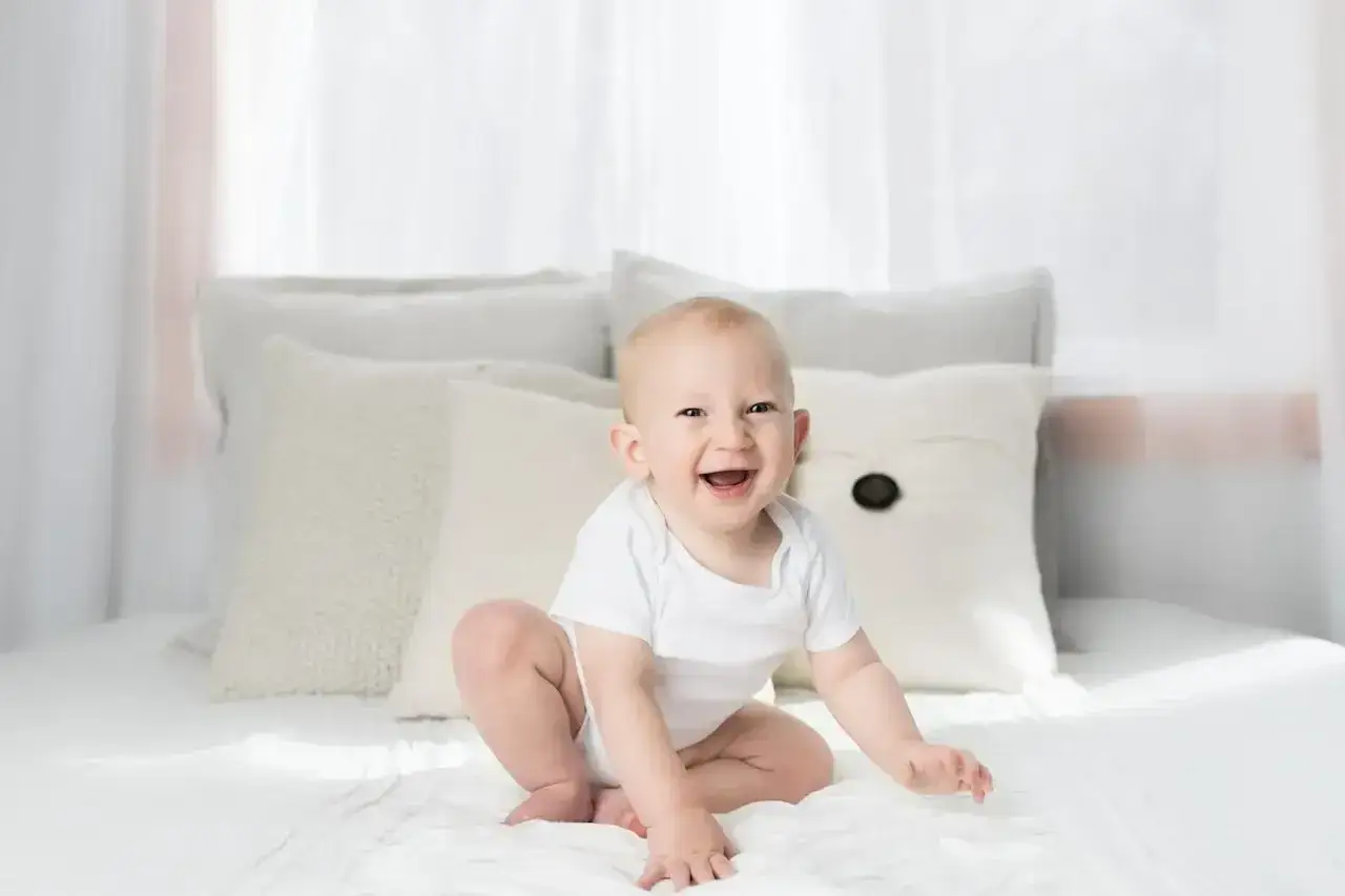 Uśmiechnięte dziecko w białym body siedzi na łóżku. To uroczy widok, który może inspirować, jak pomóc dziecku w nauce siadania.