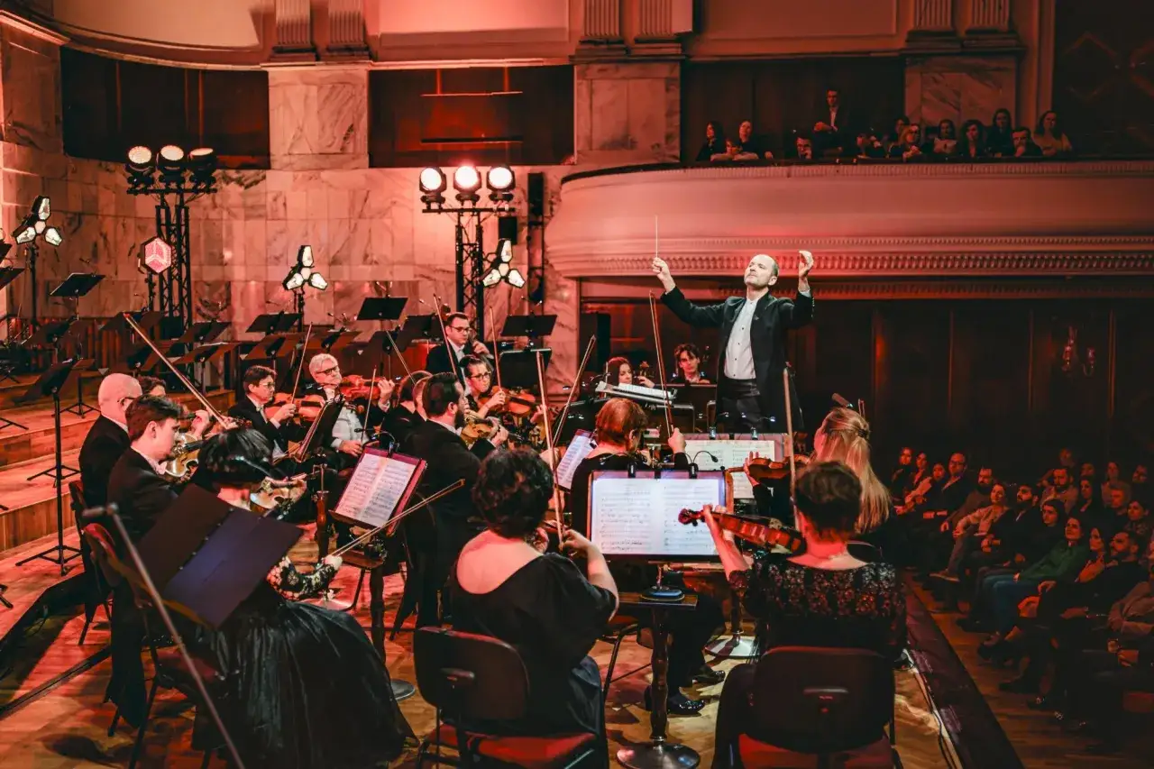 Dyrygent prowadzi orkiestrę filharmoniczną. Koncert trwa, a widzowie na balkonie wsłuchują się w muzykę.