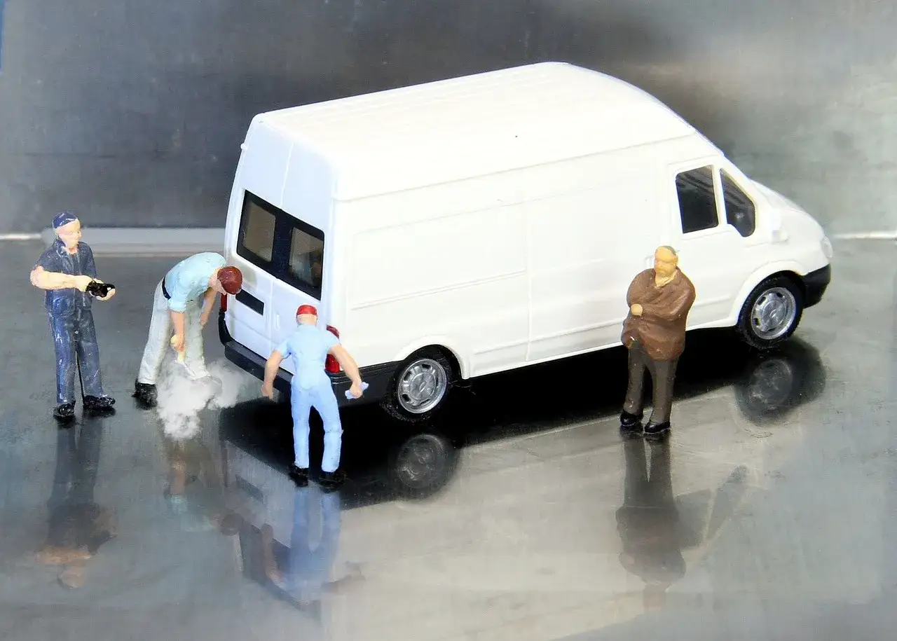 Miniaturowi pracownicy zajmują się białym busem budowlanym. Jeden z nich czyści tył pojazdu, inny trzyma narzędzie, a trzeci obserwuje.