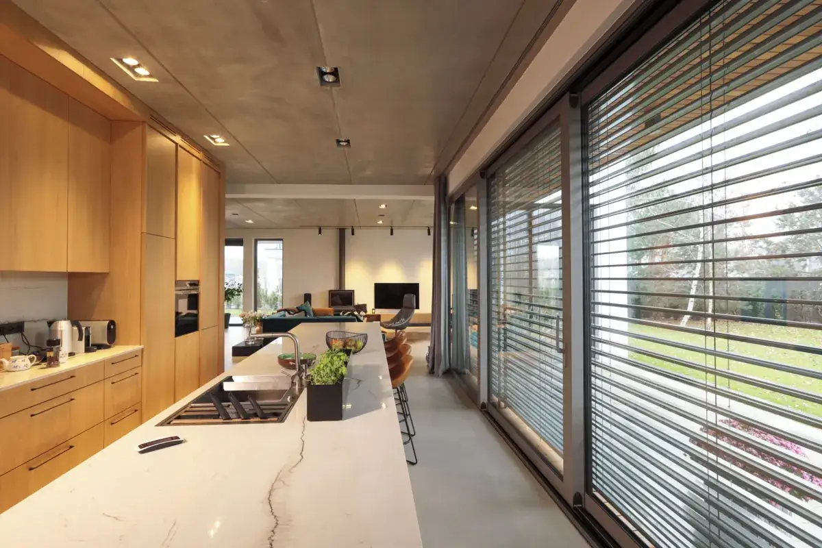 Nowoczesna kuchnia z wyspą i drewnianymi frontami, za którą widać salon. Duże okna z żaluzjami fasadowymi wewnętrznymi zapewniają prywatność.