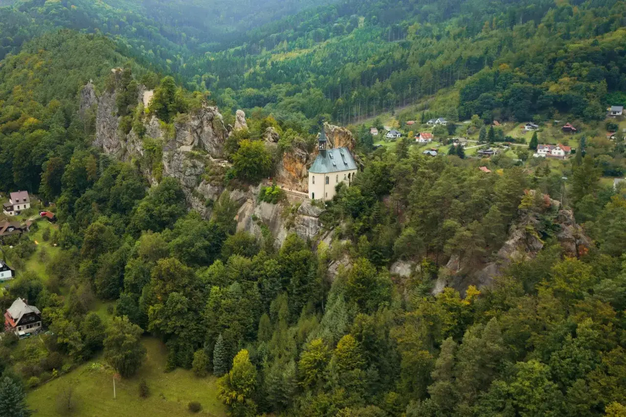 Malowniczy kościółek na skale, otoczony lasami, to jedno z **ciekawych miejsc w Czechach**. W oddali widać domki w dolinie.