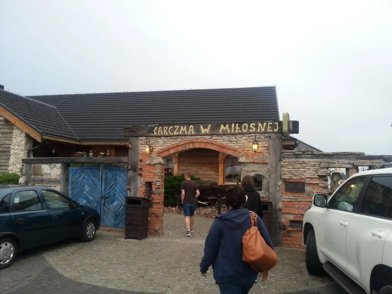 Karczma w Miłośnej, jedna z restauracji na trasie, zaprasza gości. Widać samochody i ludzi wchodzących do środka.