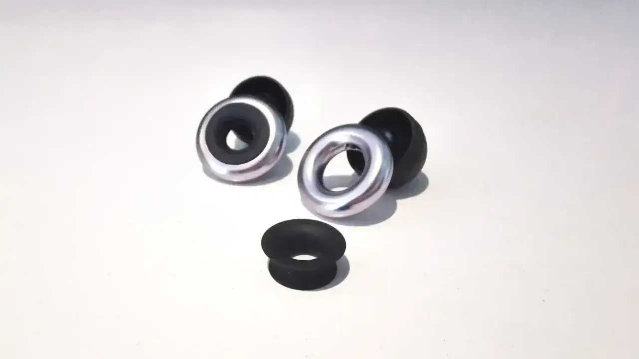 Dwa czarne, gumowe słuchawki wyglądające jak stopery, z metalowymi pierścieniami. Obok leży pojedynczy, czarny stoper.