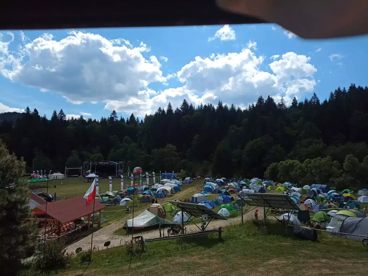 Pole namiotowe na festiwalu w Solinie. Tysiące namiotów pod błękitnym niebem z chmurami, w tle las i scena festiwalowa.