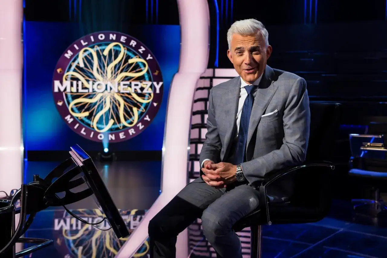 Hubert Urbański w garniturze siedzi na tle logo "Milionerzy". Zastanawia się, ile waży koszyk z pytaniami.