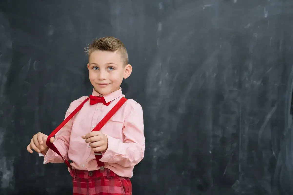 Chłopiec w różowej koszuli i czerwonych szelkach przed tablicą. Tak jak powinno czytać dziecko w 2 klasie, z uśmiechem i pewnością siebie.