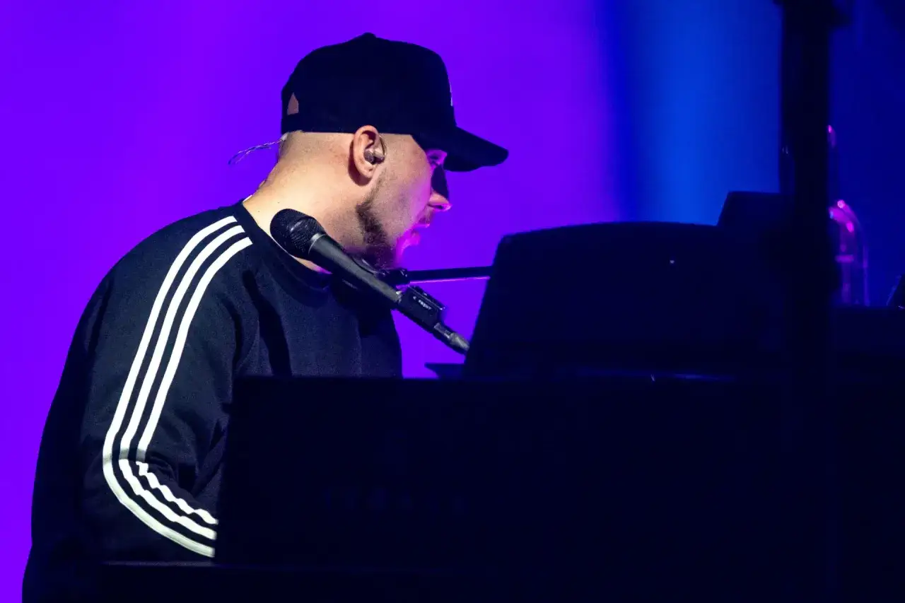 Artysta w czapce z daszkiem gra na pianinie. Koncert Korteza trwa, a fioletowe światło podkreśla atmosferę.