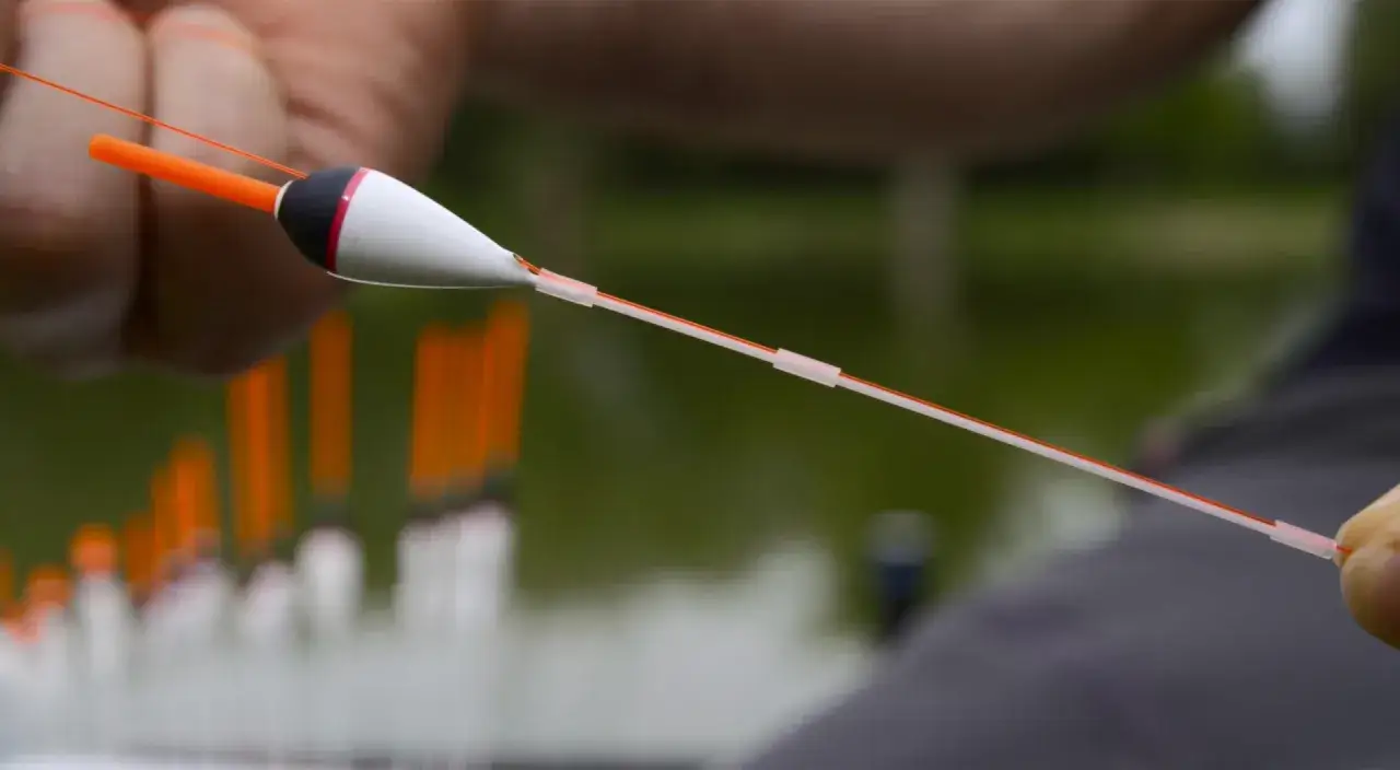 Zbliżenie na rękę trzymającą spławik wędkarski z pomarańczową antenką i biało-czarnym korpusem.