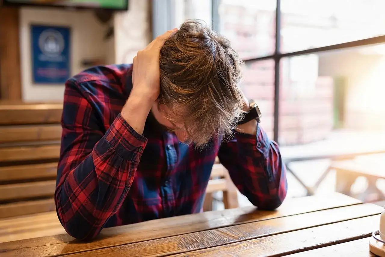 Mężczyzna w kratę z rękami na głowie siedzi przy drewnianym stole.