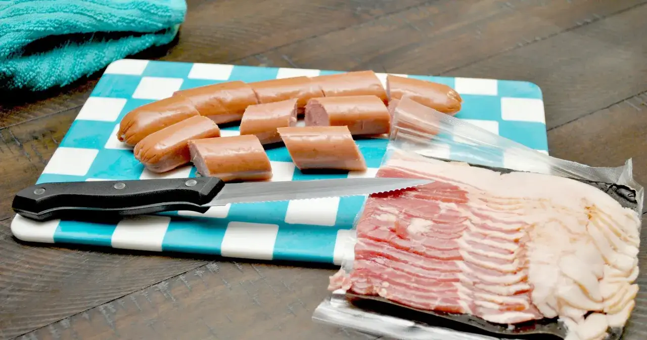 Kiełbaski pokrojone na desce w kratkę, obok plastry boczku. Zastanawiasz się, ile gram mięsa na osobę?