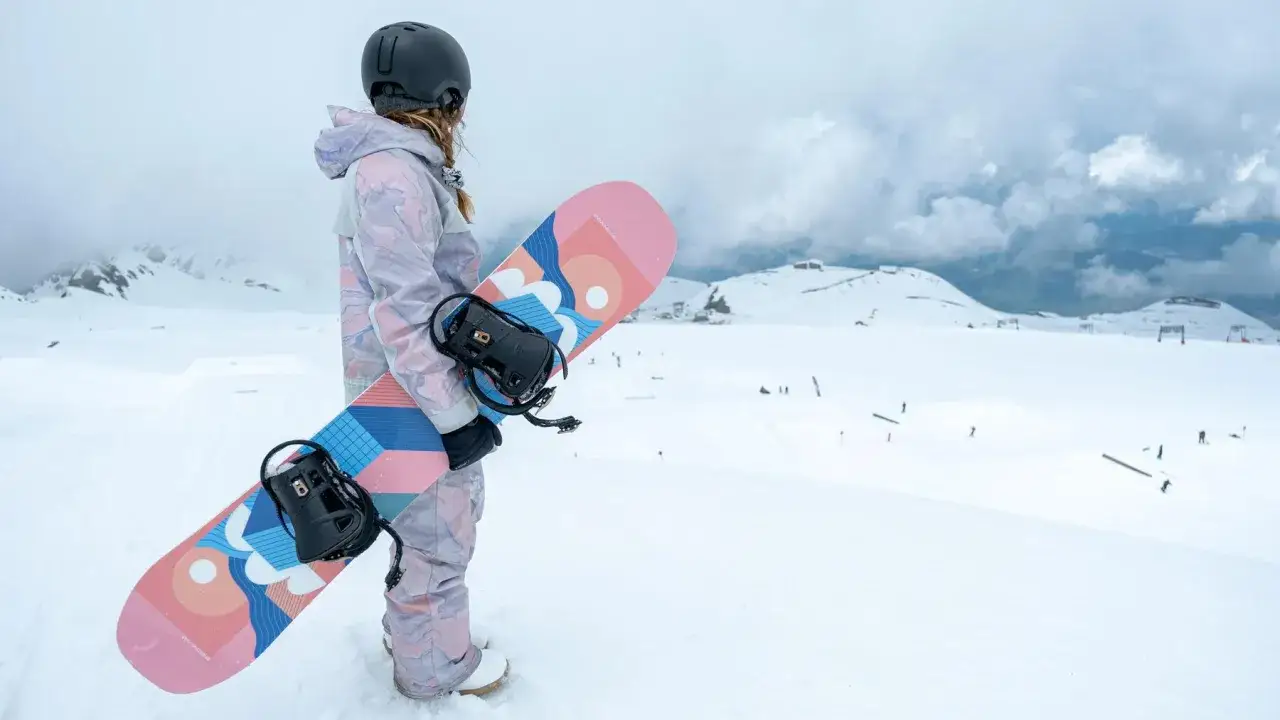 Snowboarder w kasku i kolorowym stroju trzyma deskę snowboardową z grafiką.