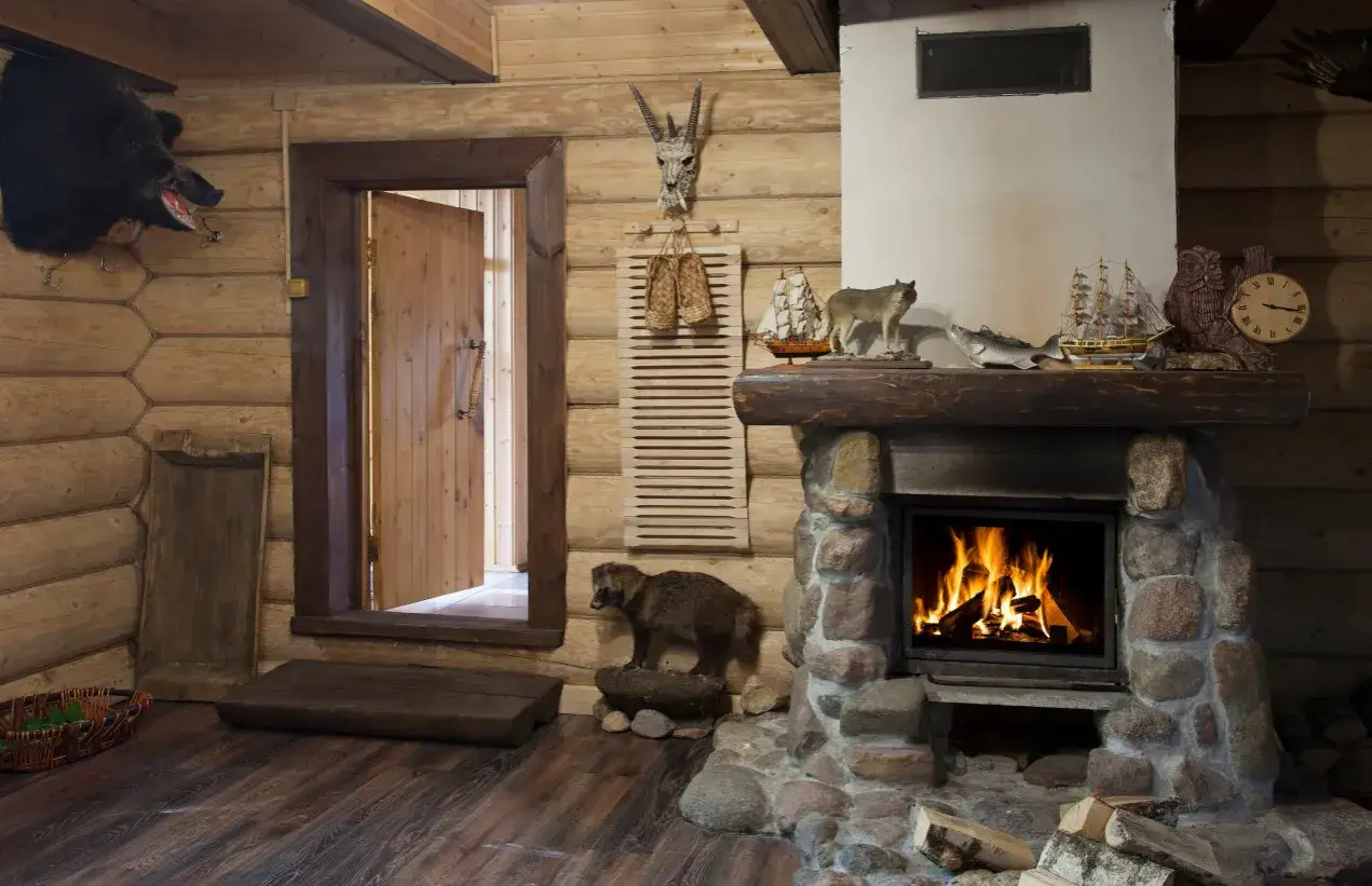 Przytulny kominek w domku drewnianym, z kamiennym wykończeniem i płonącym ogniem. Na półce nad nim stoją figurki zwierząt i statków.