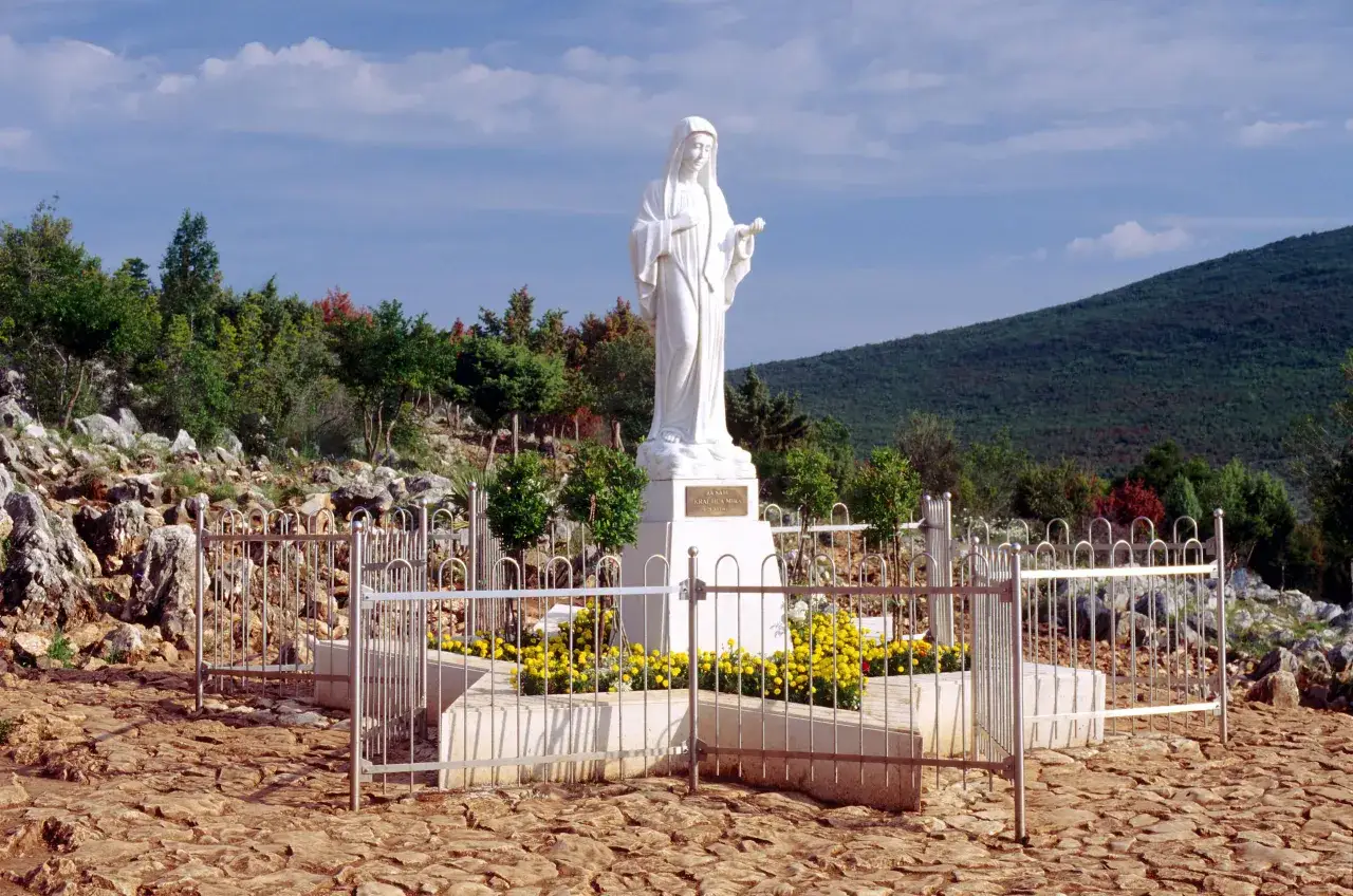 Biały posąg Matki Bożej na wzgórzu, otoczony kamieniami i ogrodzeniem. Pytanie "czy w Medjugorie jest bezpiecznie" nabiera tu szczególnego znaczenia.