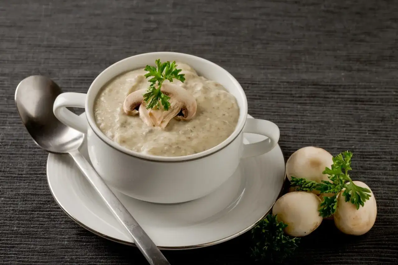 Aksamitny sos pieczarkowy w białej filiżance, ozdobiony plasterkiem pieczarki i natką pietruszki. Obok leżą świeże pieczarki.