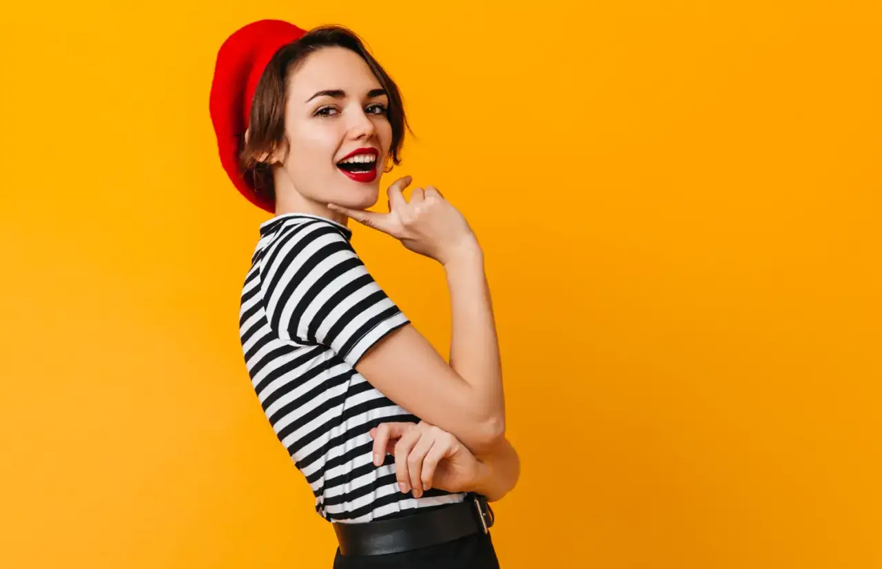 Kobieta w czerwonym beretce i pasiastym sweterku, z ustami pomalowanymi na czerwono, uśmiecha się na tle pomarańczowym. To kwintesencja paryskich kolorów.