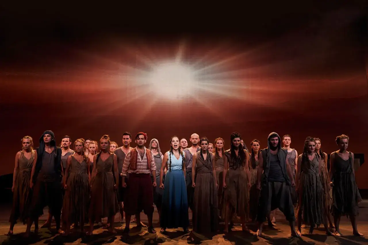 Aktorzy w teatrze roma w strojach ludowych, na tle dramatycznego, pomarańczowego nieba.