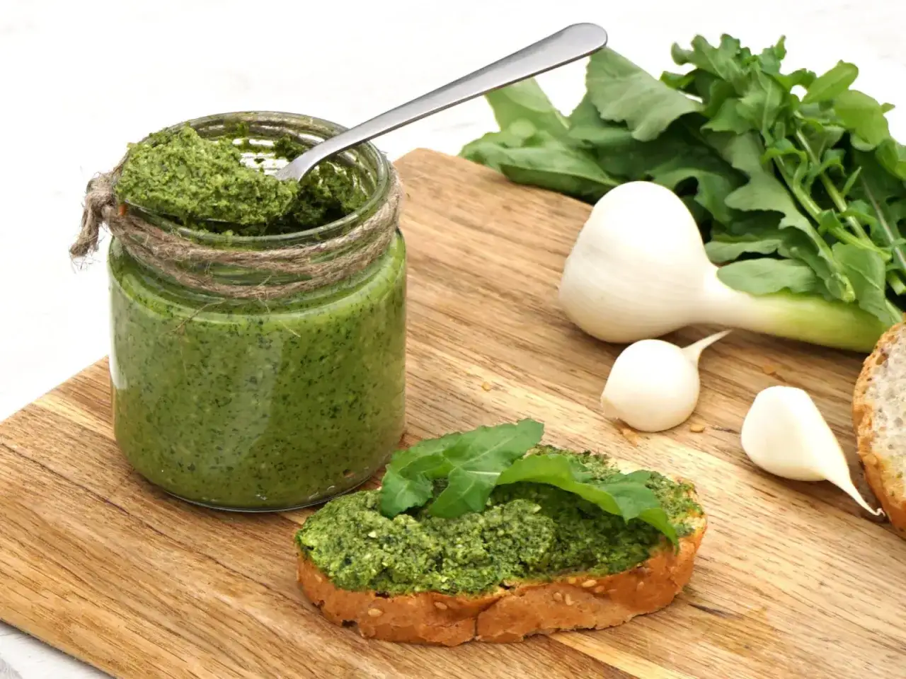 Słoik domowego pesto z rukoli, łyżka w środku, kromka chleba z pastą i świeże składniki: czosnek i rukola.