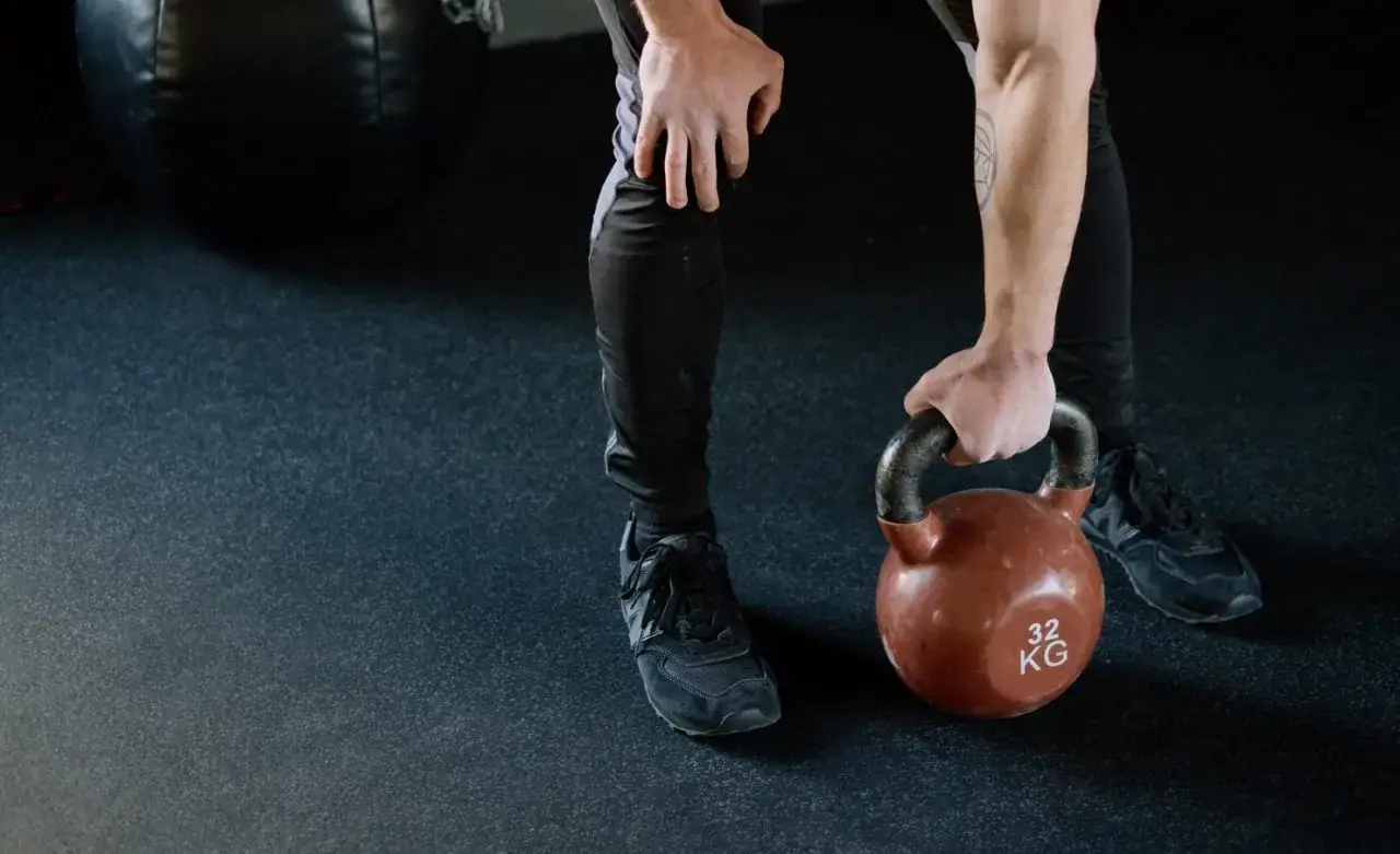 Mężczyzna w czarnych legginsach i butach sportowych podnosi kettlebell 32 kg. Zastanawia się, czy po masażu można iść na trening.
