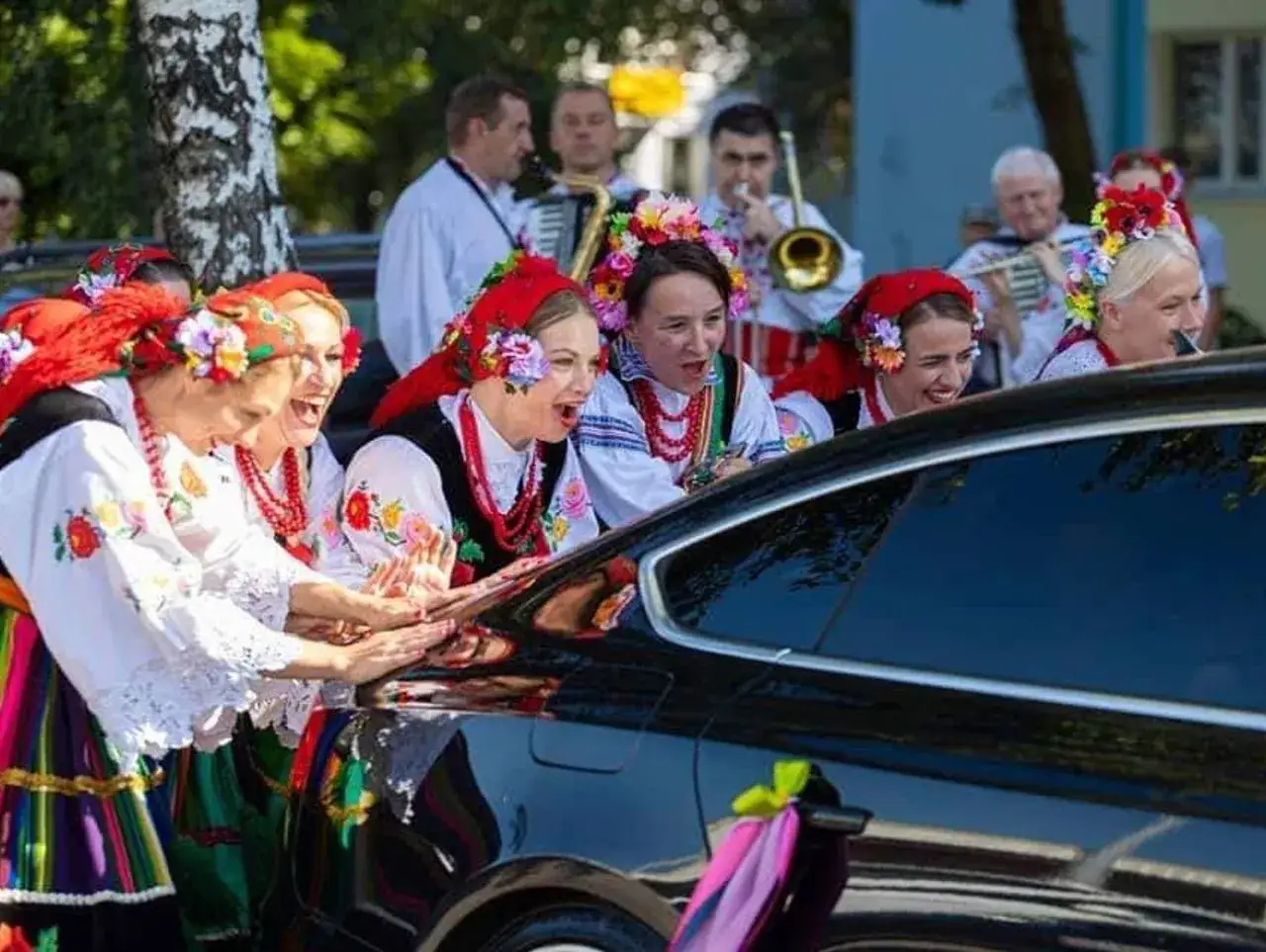 Kobiety w strojach ludowych śpiewają, jak w tekście piosenki "Gdzie ci mężczyźni", przy czarnym samochodzie ozdobionym wstążkami.
