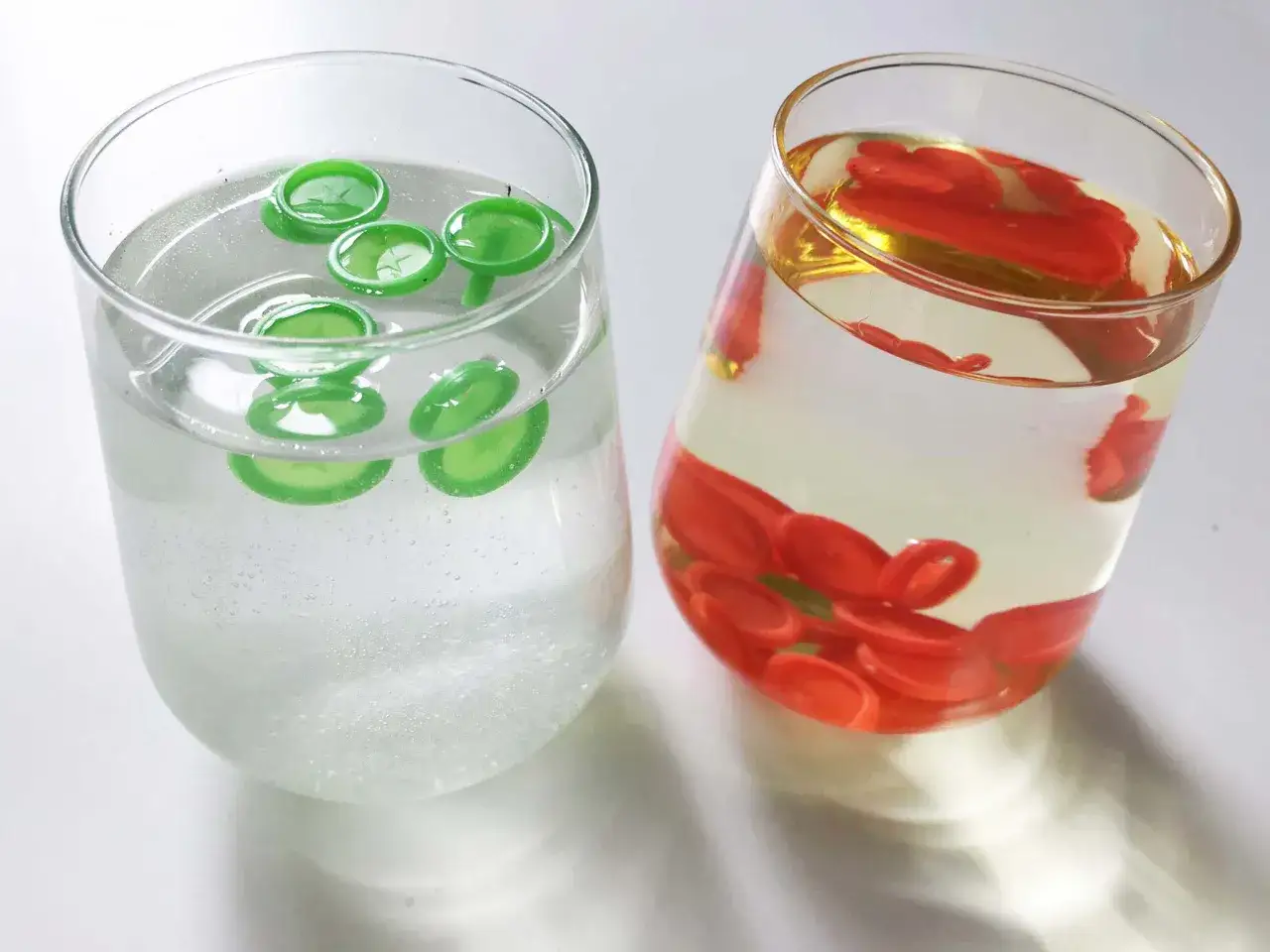 Dwa szklane kieliszki z wodą i kolorowymi, okrągłymi przedmiotami.