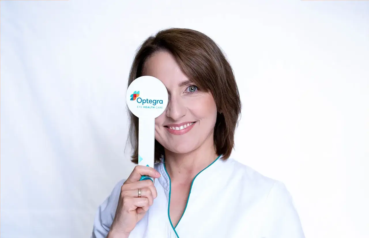 Uśmiechnięta kobieta w białym fartuchu zakrywa jedno oko tabliczką z logo "Optegra Eye Health Care", doświadczając chwilowej utraty ostrości widzenia podczas badania.