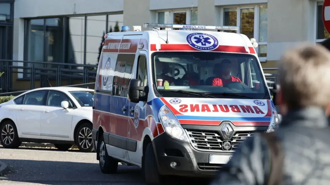 Ambulans z ratownikami medycznymi na tle budynku i białego samochodu.
