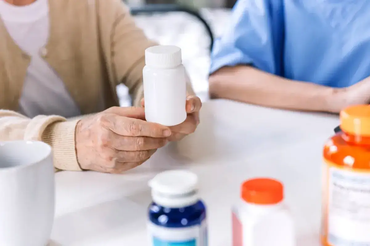 Dłoń starszej osoby trzyma białą buteleczkę leku. Na stole stoją inne opakowania leków.