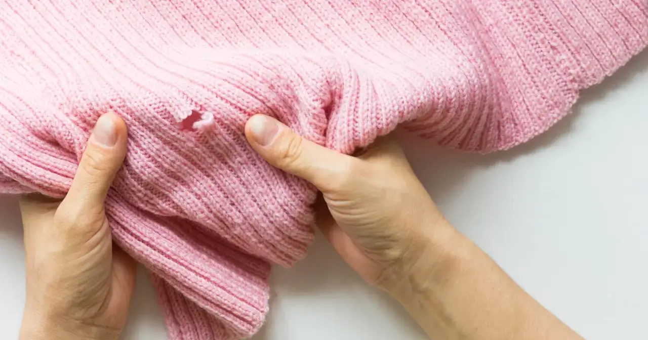 Dłonie trzymają różowy sweter z dziurą, pokazując problem, jak zaszyć dziurę w dresach.