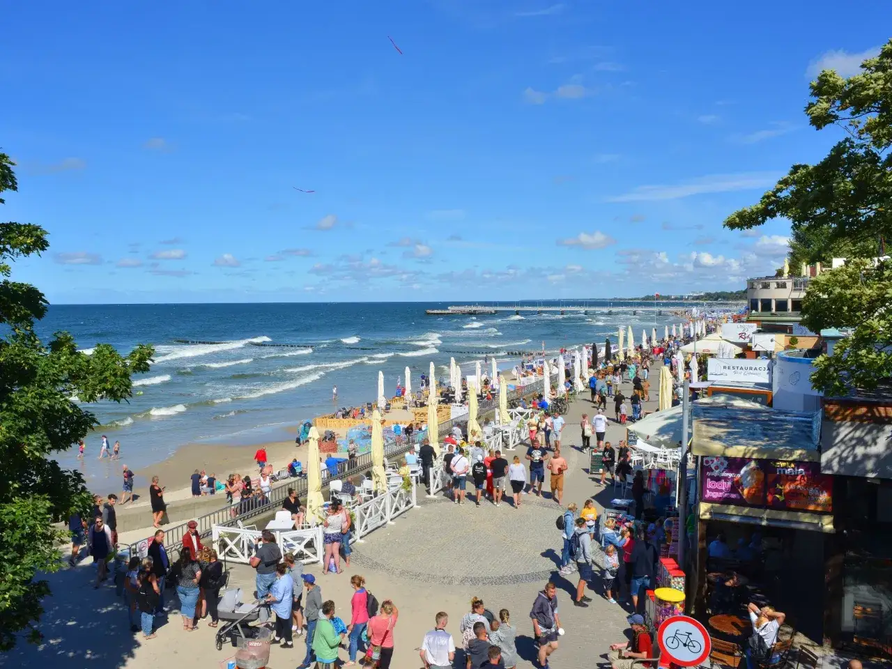 Tłum ludzi spaceruje promenadą w Kołobrzegu, obok plaży i restauracji. Zastanawiasz się, ile kosztuje klimatyczne w Kołobrzegu?