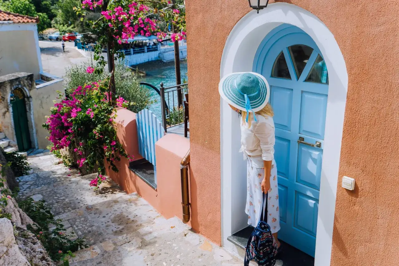 Kobieta w kapeluszu przy niebieskich drzwiach, gotowa na wakacje do Grecji all inclusive. W tle kwitnące bugenwille i morze.