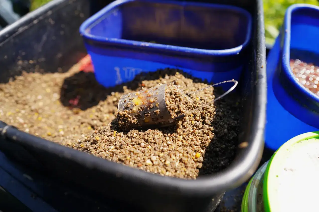 Przezroczysty koszyczek z zanętą feeder bait w czarnym wiadrze, gotowy do łowienia.