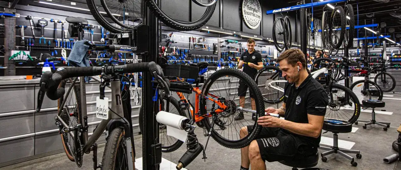 W warsztacie fabryki rowerów mechanik naprawia rower. W tle wiszą rowery i narzędzia.