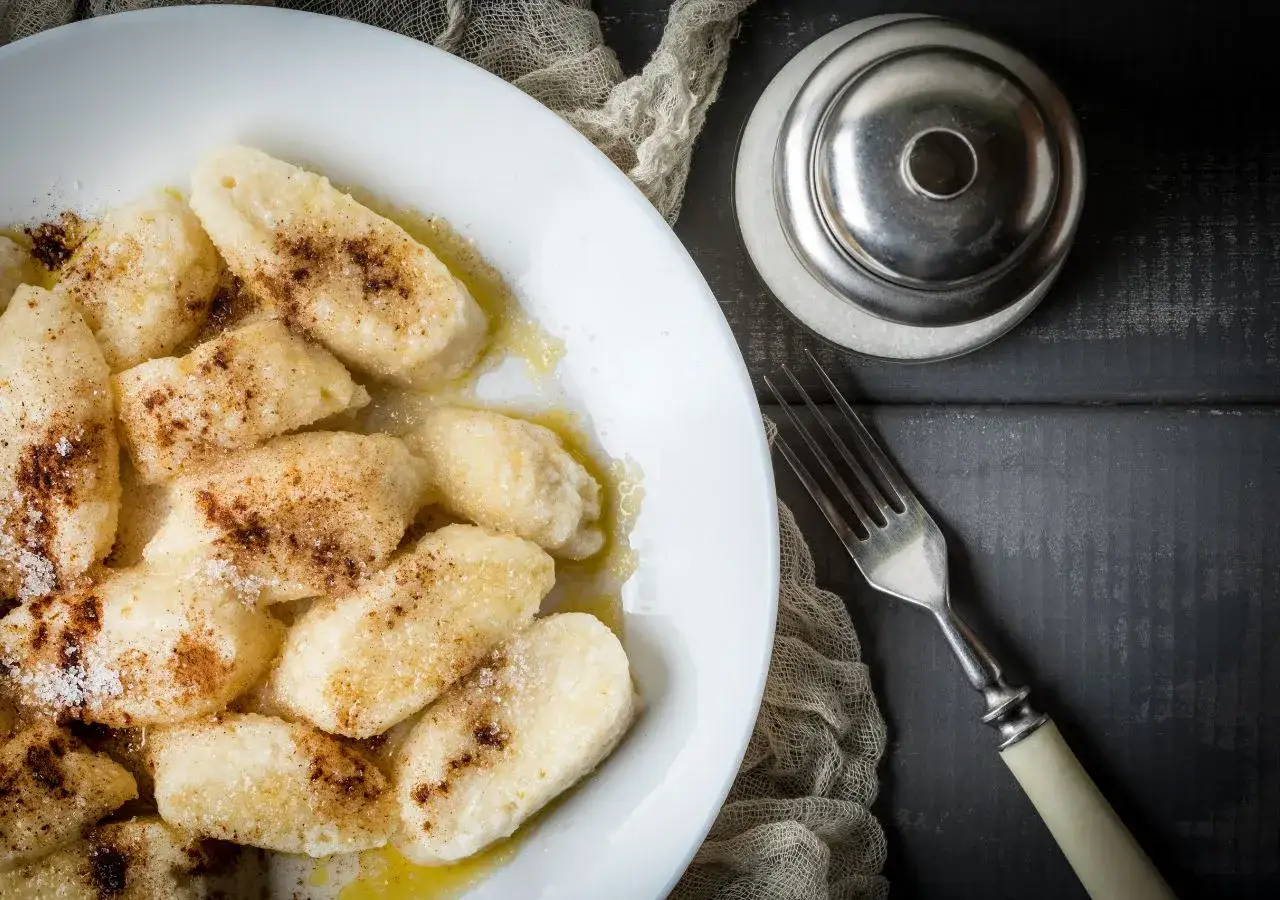 Kluski śląskie na białym talerzu, posypane cukrem i cynamonem, obok widelec i cukiernica.