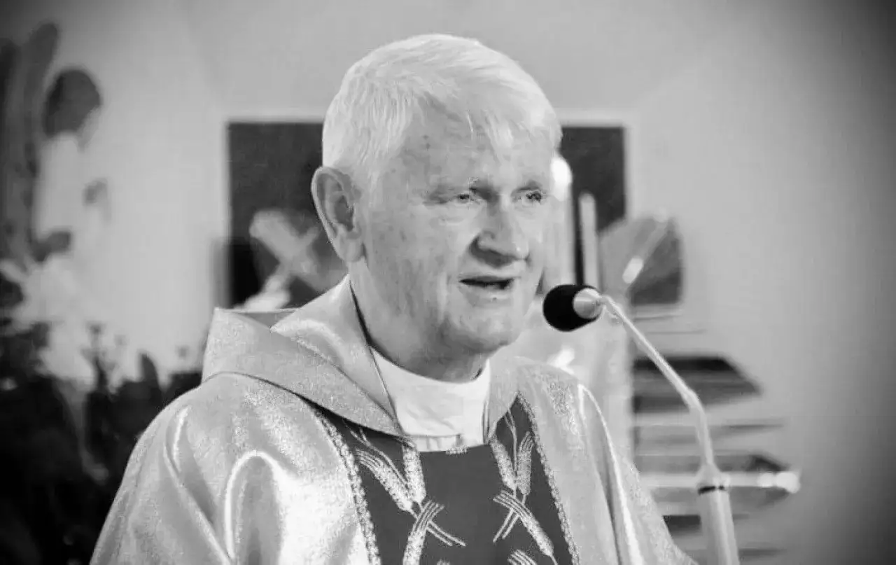 Czarno-białe zdjęcie starszego księdza w szatach liturgicznych, mówiącego do mikrofonu.