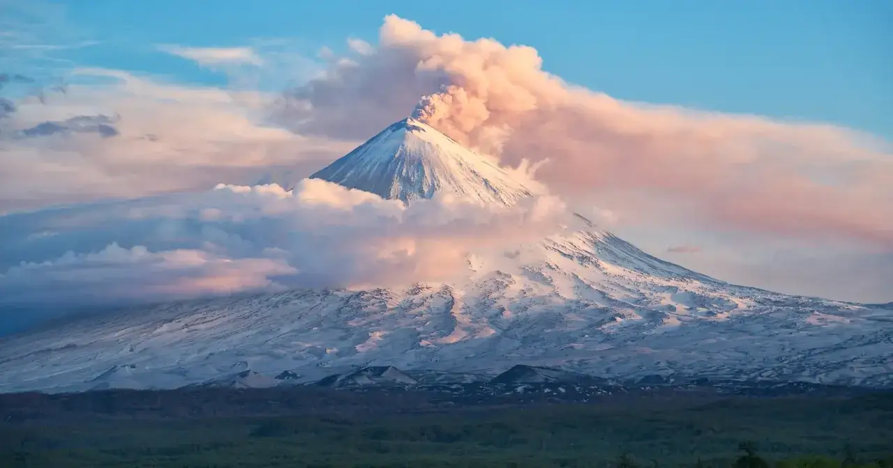Śnieżny stożek wulkanu na Kamczatce wyrzuca dym i popiół w różowe chmury o zachodzie słońca.