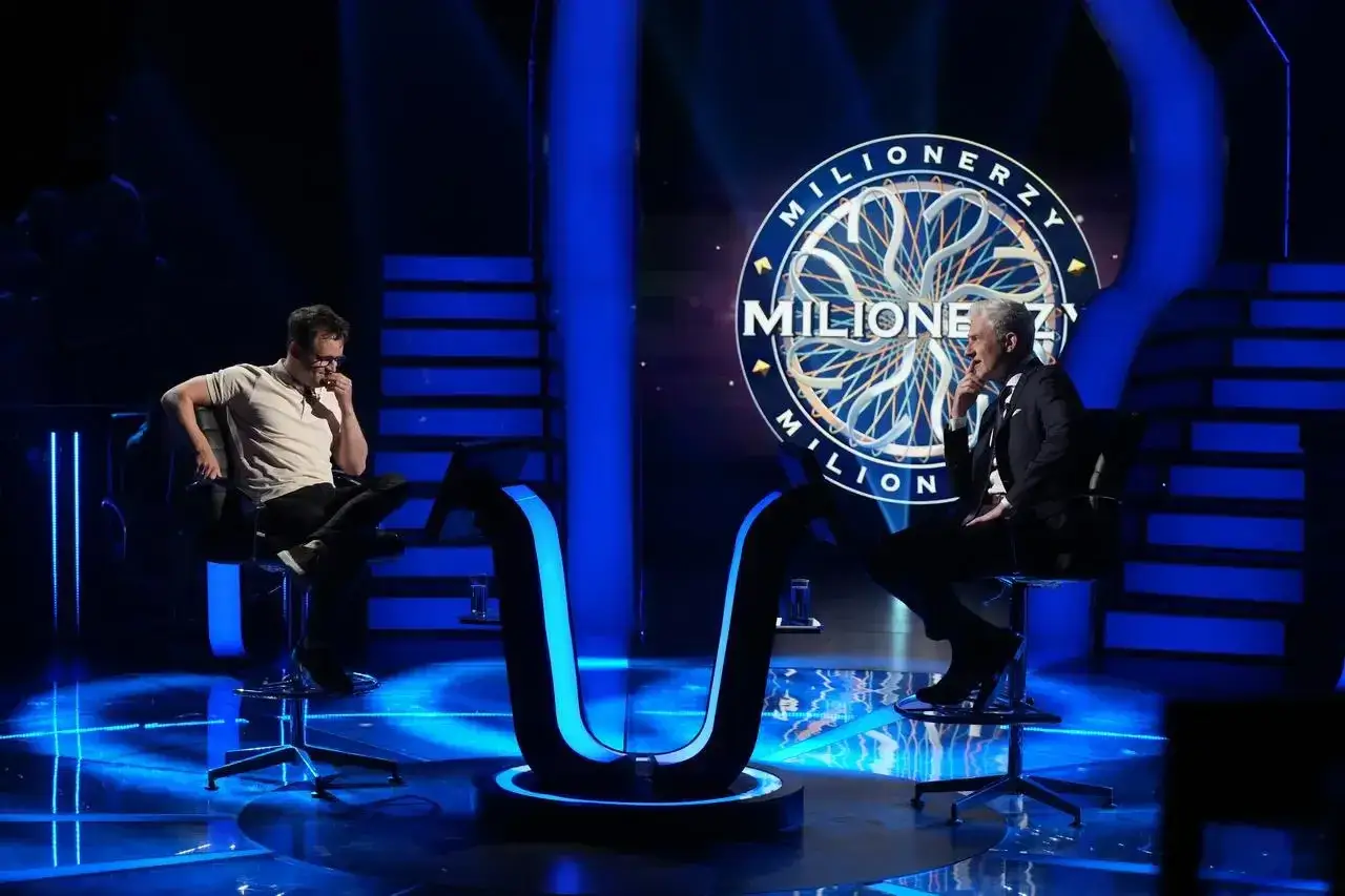 Dwóch mężczyzn siedzi naprzeciwko siebie na scenie "Milionerzy". Jeden w białej koszulce, drugi w garniturze.