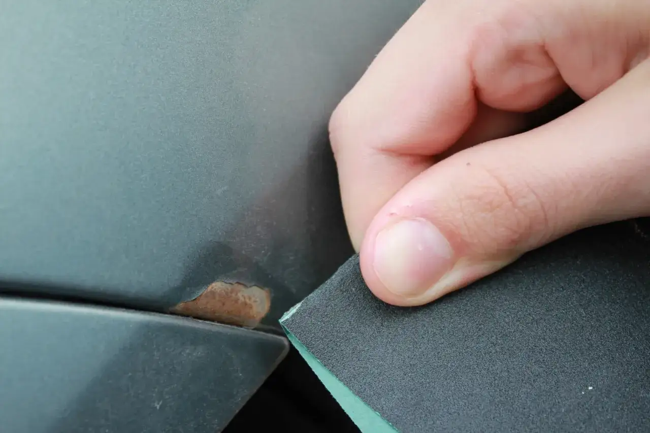 Dłoń z papierem ściernym przygotowuje fragment karoserii do naprawy, pokazując jak zrobić zaprawkę lakierniczą na zardzewiałym miejscu.