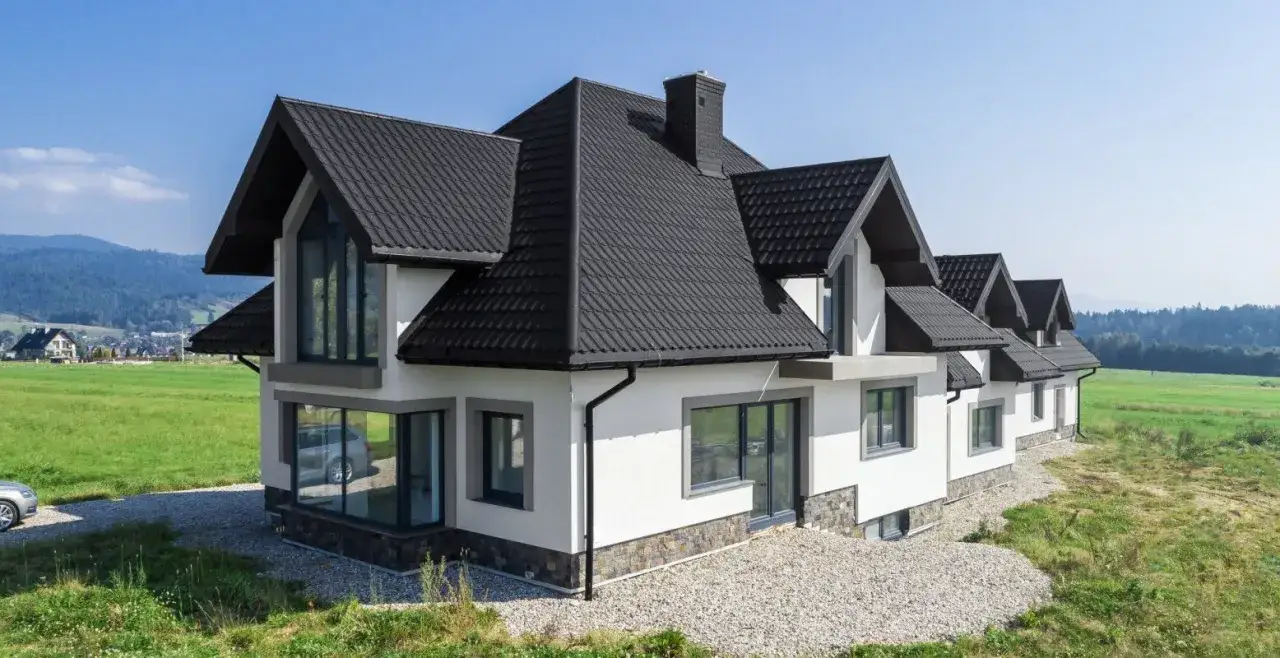 Nowoczesny dom z czarnym, wielospadowym dachem, otoczony zielenią i górami.